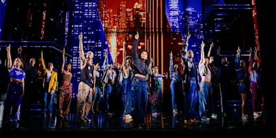 A diverse group of performers stands on stage with one arm raised, set against a colorful cityscape backdrop with bright lights and tall buildings.