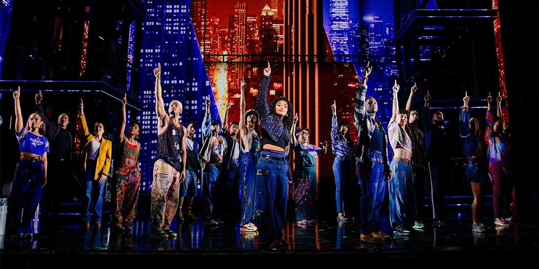 A diverse group of performers stands on stage with one arm raised, set against a colorful cityscape backdrop with bright lights and tall buildings.