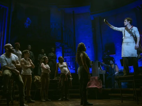 A group of actors on stage in a dark, blue-lit theater scene, attentively watching a person in a white apron holding up a cup.