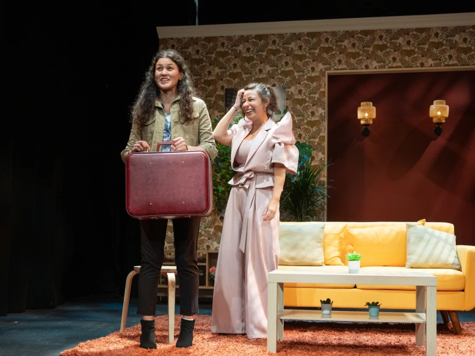 Two women on stage, smiling. One holds a red suitcase, and the other is adjusting her hair. They're in a living room set with retro wallpaper, a yellow couch, and small table with plants.