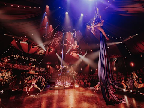 Aerial performers and acrobats entertain a seated audience under dramatic lighting in a circus tent labeled “The Five Tops.”.
