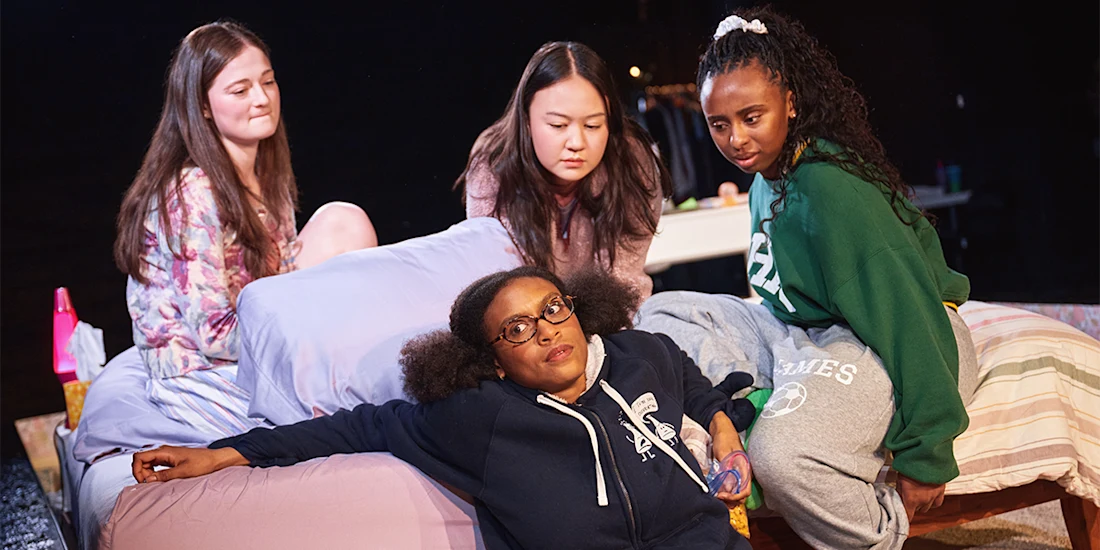 Four young women sit and lay on and around a bed, appearing to have a serious conversation in a dimly lit room.