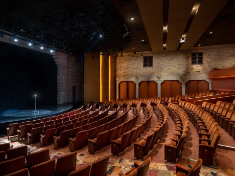 An empty theater with rows of brown seats facing a stage lit by a single spotlight, surrounded by brick walls and overhead lighting.