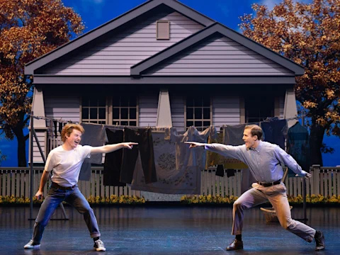 Two men enthusiastically point at each other while performing on stage in front of a house facade and a clothesline with laundry.