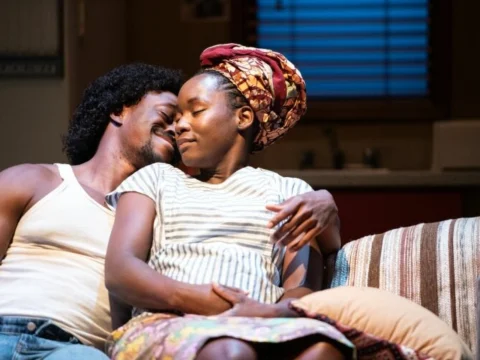 Two people sit closely together on a couch. One person rests their head on the other's shoulder, eyes closed and smiling gently. The person wearing a striped shirt and headwrap appears relaxed and content.