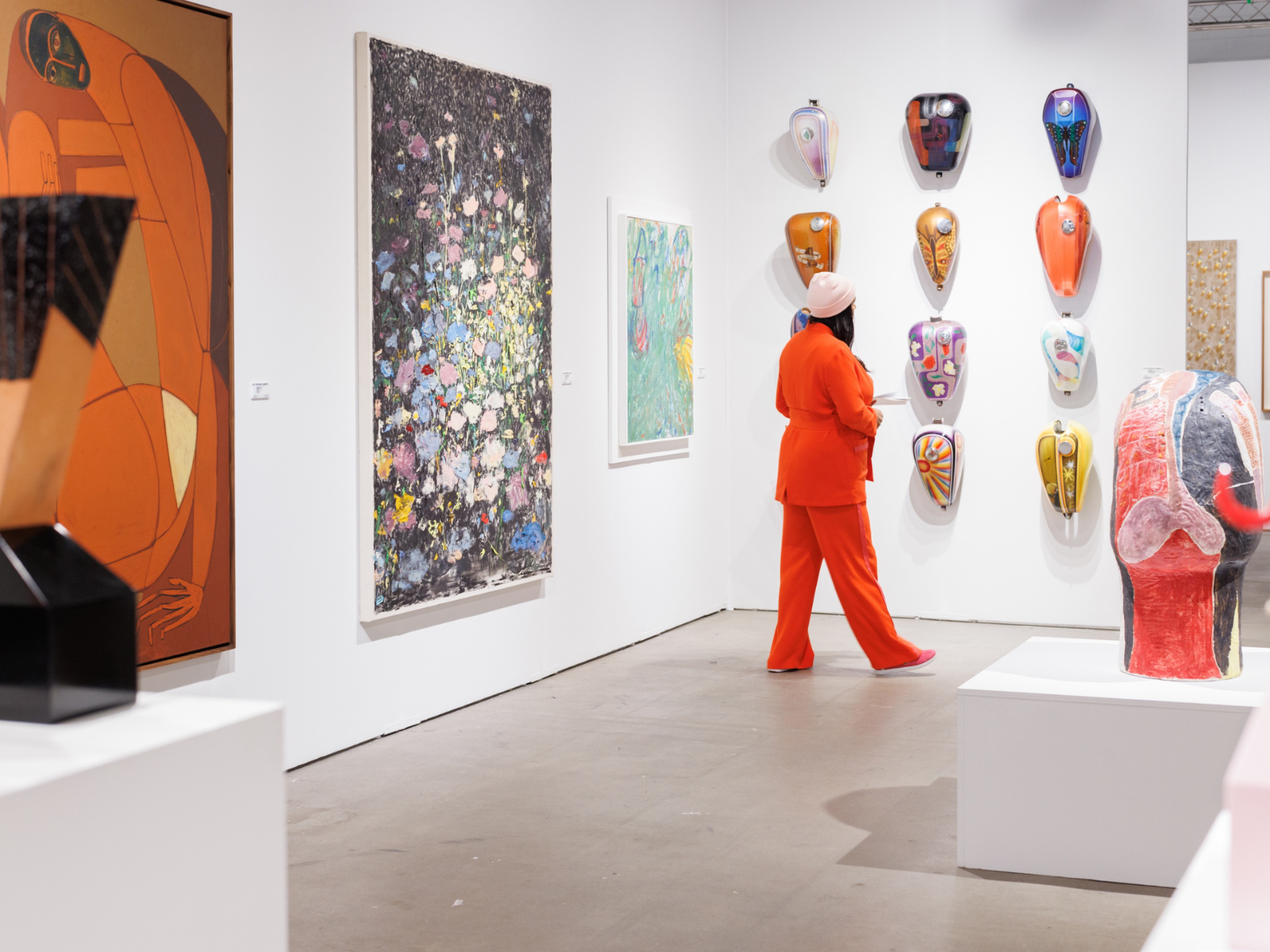 A person dressed in orange stands in an art gallery, viewing colorful mask-like sculptures displayed on a white wall among various paintings and sculptures.