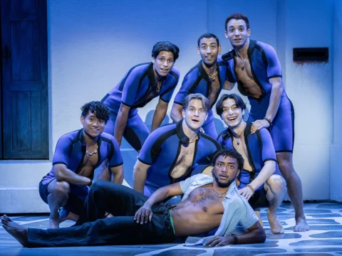 A group of six individuals in matching purple and black wetsuits pose energetically, with one person lounging in front on the ground, shirtless, against a blue and white backdrop.