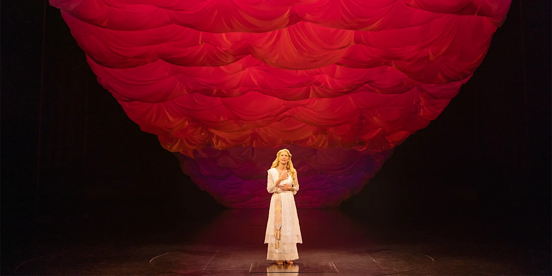 A woman in a long, light-colored dress stands alone on stage beneath a dramatic ceiling of layered red fabric.