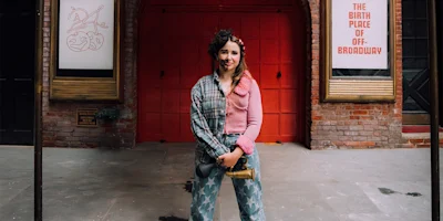 A person dressed in eclectic clothing stands on a city sidewalk in front of a red door and signs reading "The Birth Place of Off-Broadway.