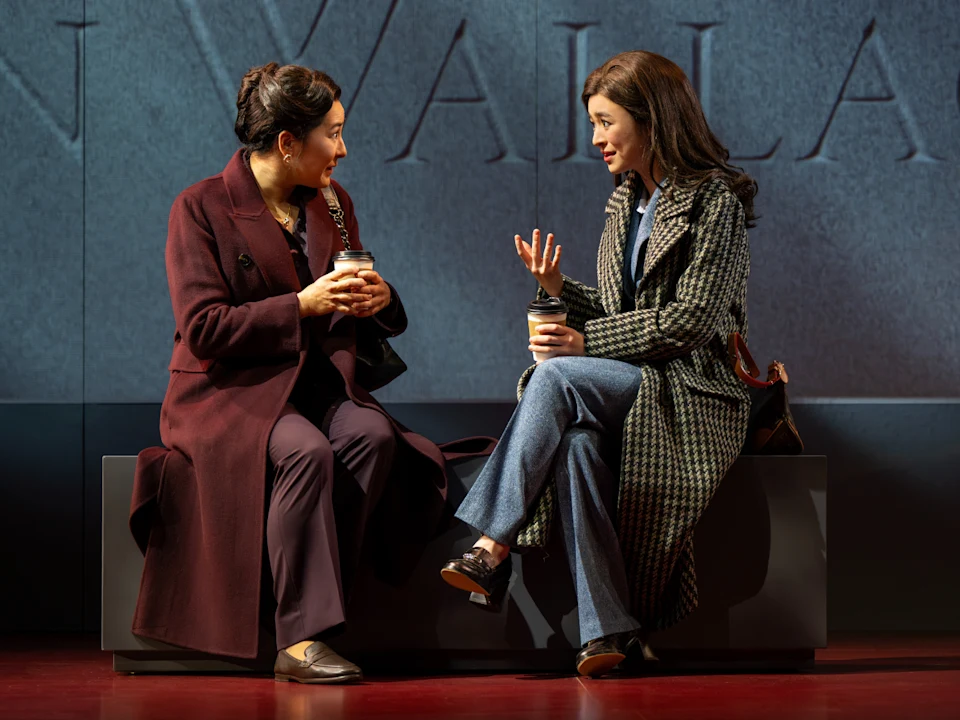 Two women in coats sit on a bench, holding coffee cups and having a conversation in front of a stone wall backdrop.