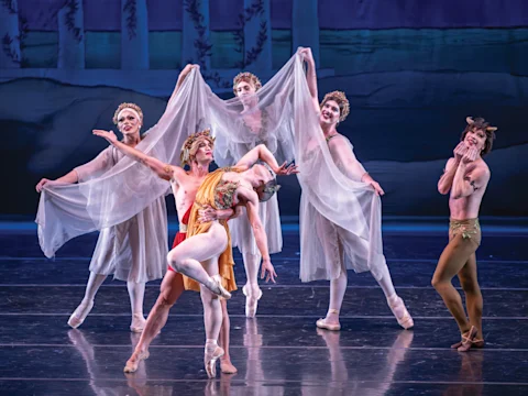 Five ballet dancers perform on stage; one male dancer in orange lifts a female dancer while three others hold sheer fabric in the background and a fifth dancer reacts dramatically on the right.