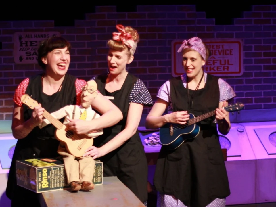 Three women in black aprons, with 1940s-style hairdos, perform on stage. One holds a puppet dressed as a baker, another plays a ukulele, and the third smiles while standing in front of a brick backdrop.