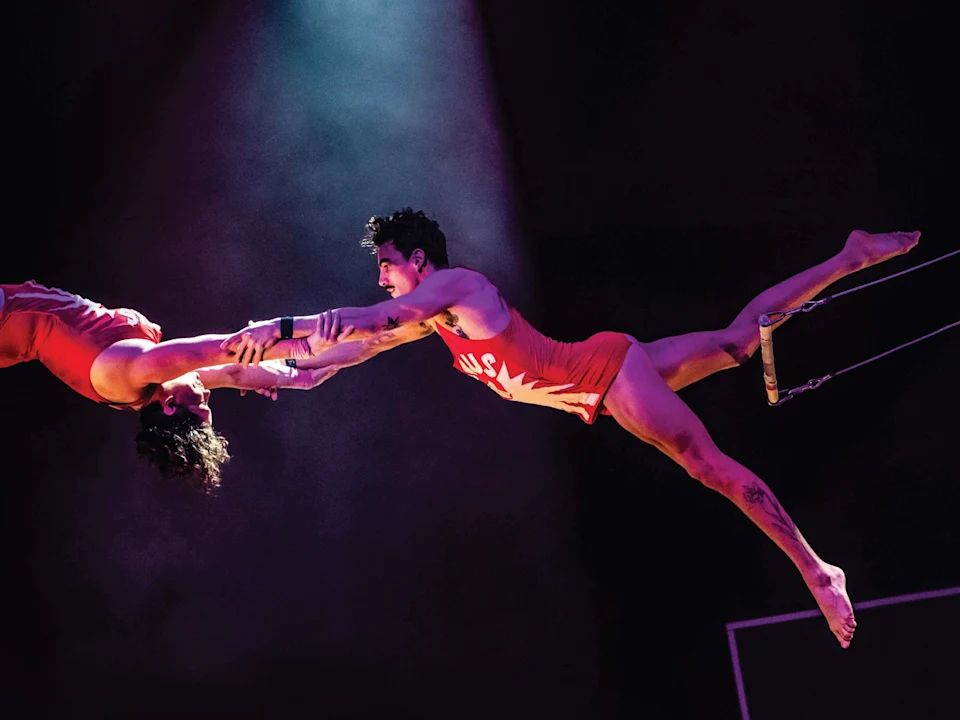 Two acrobats in red costumes perform a mid-air trapeze act, reaching out to grasp each other's hands under a spotlight.