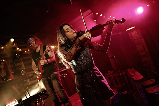 A woman plays the violin on stage under pink and purple lights, with a guitarist and keyboardist performing in the background.