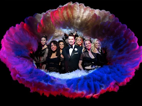 Production photo of Chicago on Broadway, showing a group of performers in formal and show costumes stand together, framed by large, colorful feather fans against a black background.