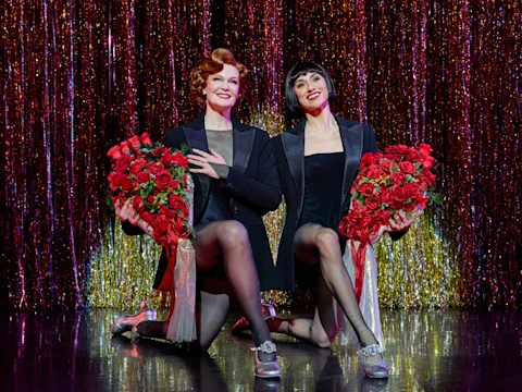 Production photo of Chicago on Broadway, showing two performers in black costumes kneel on stage holding large bouquets of red roses, with a glittery red and gold curtain backdrop.