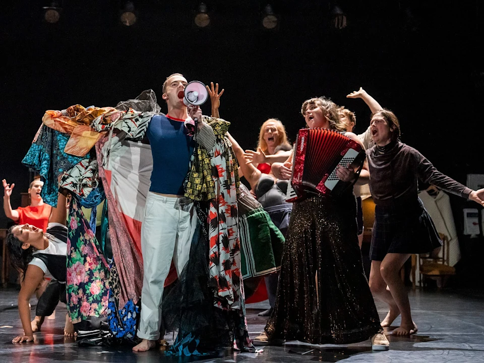 A group of performers on stage, one using a megaphone and draped in colorful fabrics, another playing an accordion, with others gesturing energetically in the background.
