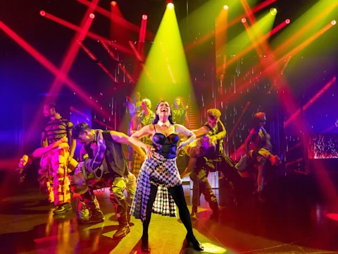 Performers dance energetically on a brightly lit stage with red and yellow spotlights, with one performer standing at the center in a dynamic pose.