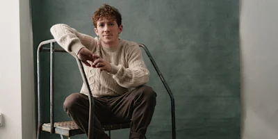 A young man with curly hair sits on a metal ladder, wearing a cream cable-knit sweater and dark pants, against a plain green background.