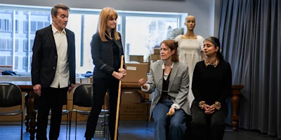 Four adults and a mannequin are in a room with boxes; two people stand, and two are seated, appearing to converse.