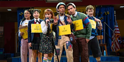 Six young people stand on stage during a spelling bee, each wearing a numbered sign around their neck and holding a microphone, with a colorful backdrop behind them.