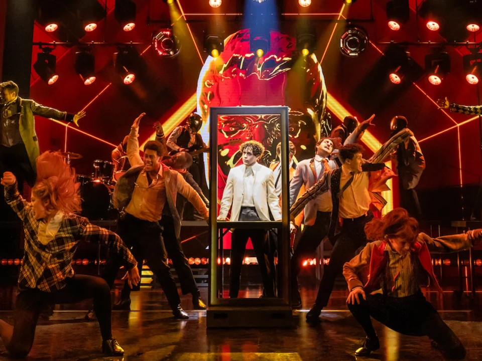 A group of performers dressed in eclectic costumes dance energetically on a stage with vibrant lighting and effects, featuring one central figure in a glass booth.