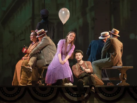 A stage scene featuring performers in period costumes. A young woman in a purple dress and holding a balloon is seated center-stage, surrounded by other actors on benches, some wearing straw hats.