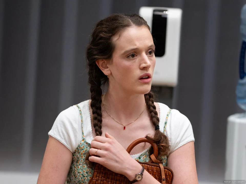 A woman with braided hair, wearing a floral dress and holding a wicker bag, stands against a background with a white and blue object visible.