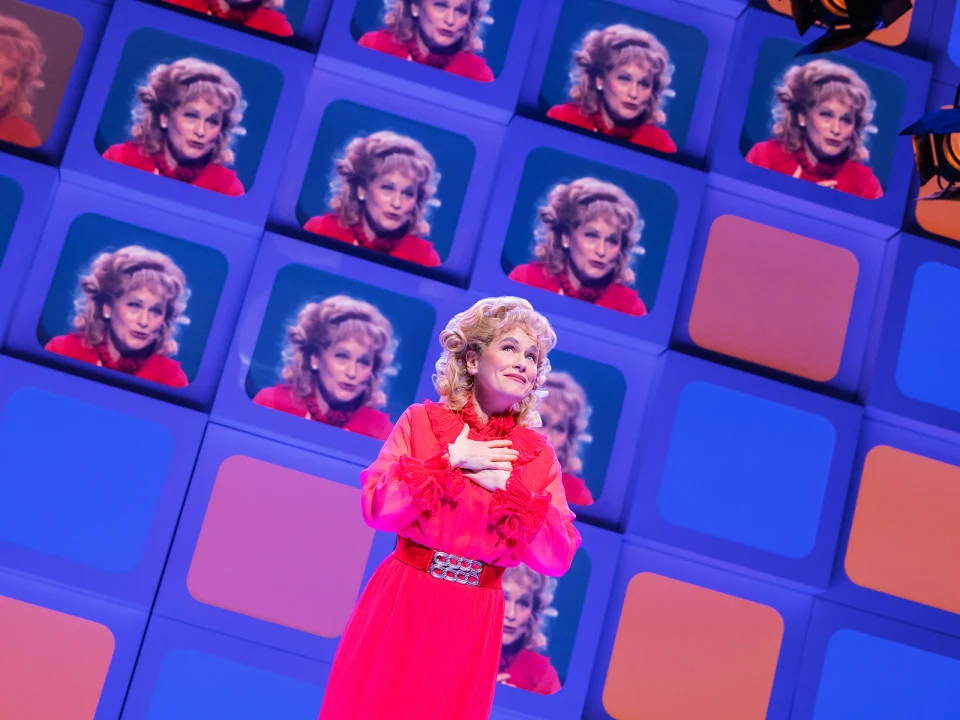 A person in a bright red outfit stands in front of a wall of screens displaying their image.