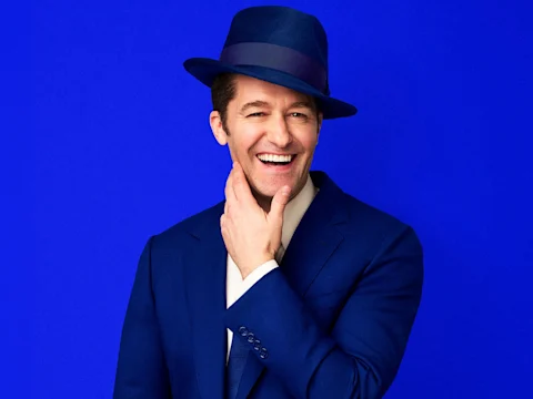 A man in a blue suit and matching hat smiles while touching his chin against a solid blue background.