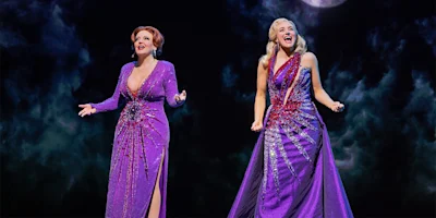 Two women in purple, glittering gowns sing on stage against a dark, cloudy background with a hint of a full moon above.