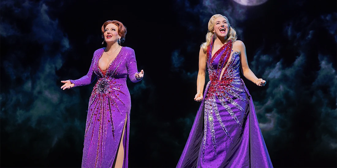 Two women in purple, glittering gowns sing on stage against a dark, cloudy background with a hint of a full moon above.