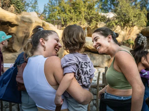 Two women smile at a young child they are holding at an outdoor location with trees and rocks in the background. Another person wearing a hat is partially visible on the left.