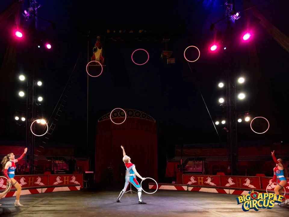 Performers in colorful costumes juggle hula hoops on a circus stage under vibrant lights.