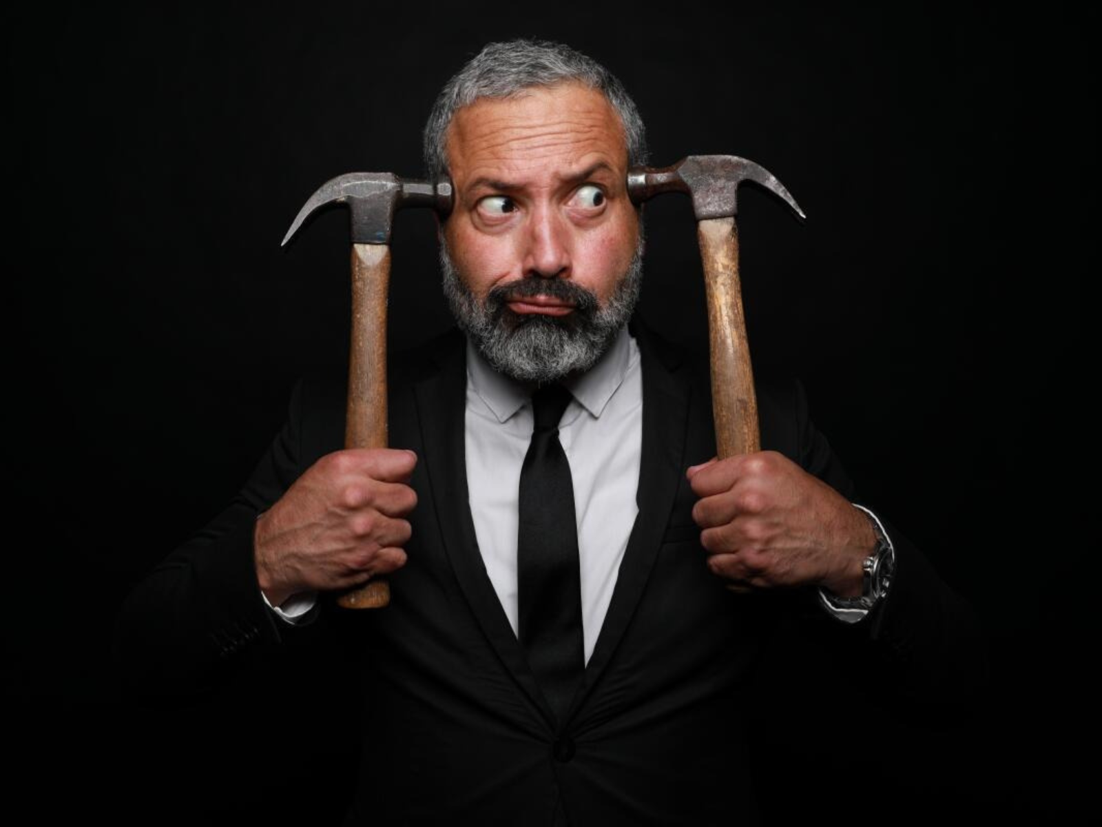 A man in a suit holds two hammers to his head, one in each hand, against a dark background, with a puzzled expression on his face.