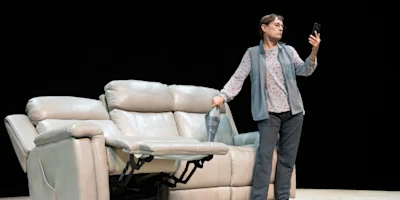 A woman stands next to a light-colored recliner sofa, holding a vacuum cleaner and looking at a smartphone.