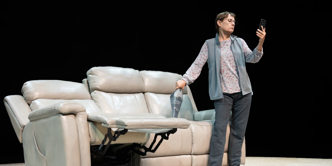 A woman stands next to a light-colored recliner sofa, holding a vacuum cleaner and looking at a smartphone.