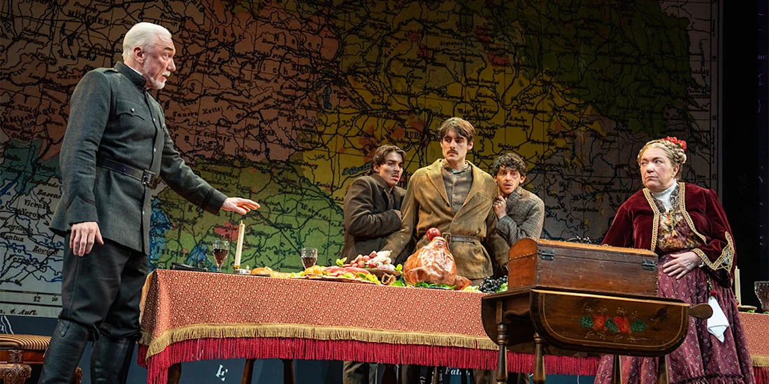 Five actors in period costumes perform a dramatic scene around a table with food, in front of a large map backdrop on stage.