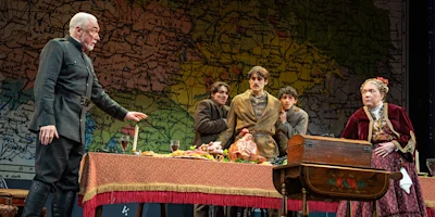 Five actors in period costumes perform a dramatic scene around a table with food, in front of a large map backdrop on stage.
