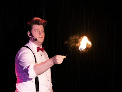 A performer in a white shirt, bowtie, and suspenders holds a stick with a flame at its tip, standing against a dark background with a headset microphone.