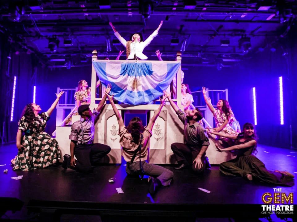 Actors perform a dramatic scene on stage with a large flag featuring blue and white stripes and a sun emblem.
