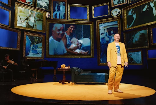 A person in a yellow outfit stands on a yellow circular stage with a blue couch, surrounded by large framed family photos and musicians in the background.