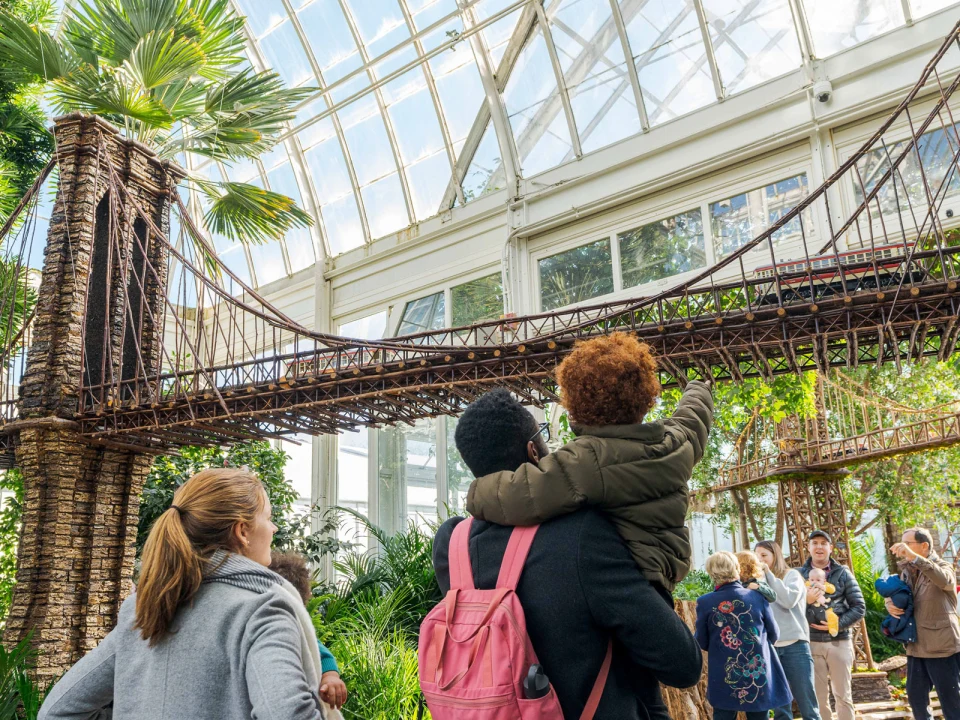 A family looks at a detailed model bridge inside a greenhouse, surrounded by plants and other visitors.