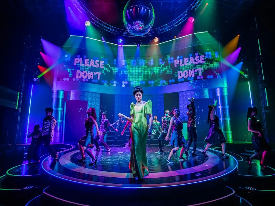 A performer in a green dress stands center stage under colorful lights, with backup dancers around and "PLEASE DON'T" displayed on screens behind them.