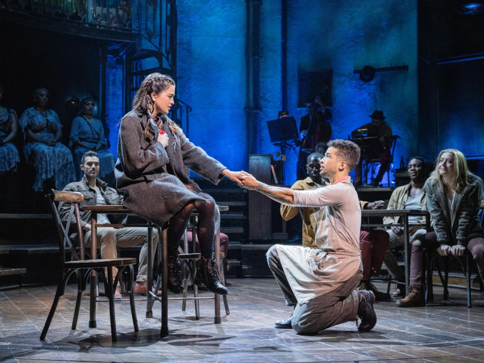 A man kneels and holds the hand of a seated woman during a stage performance, with other cast members and musicians in the background.