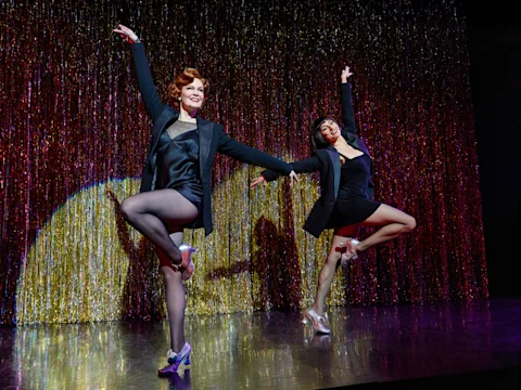 Production photo of Chicago on Broadway, showing two dancers in dark costumes and heels perform a synchronized routine on stage in front of a shimmering, metallic curtain.