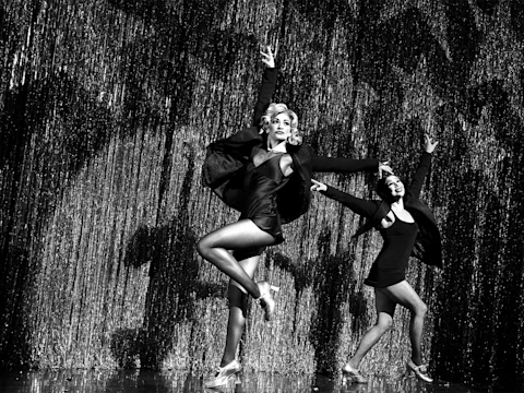 Two performers in stage costumes dance energetically in front of a glittering curtain, with one dancer in the foreground and the other slightly behind.