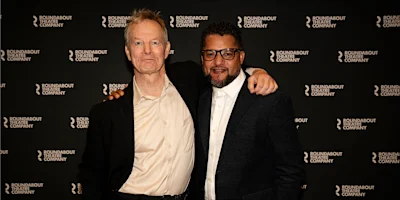 Two men in suits posing together in front of a Roundabout Theatre Company step and repeat banner.