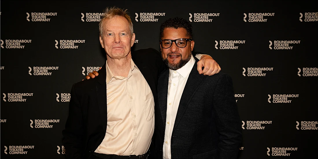 Two men in suits posing together in front of a Roundabout Theatre Company step and repeat banner.