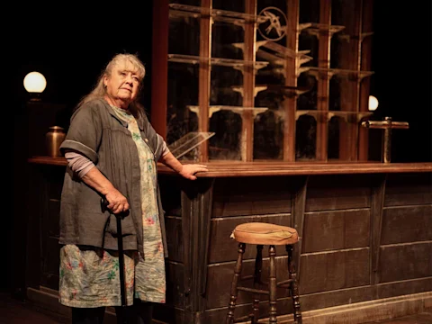 An older woman with gray hair, holding a cane, stands by a wooden bar with shelves and a stool, on a dimly lit stage.